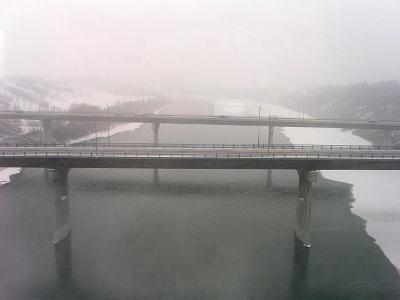 [Midway across the river looking at two bridges across the river. The snow is piled up on the water mostly on the right, so that side is probably partially iced. The background of the image is mostly a whiteout scene due to the snow.]